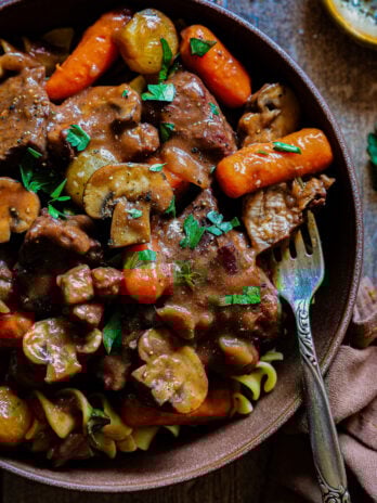 Beef bourguignon served over egg noodles with carrots, mushrooms, pearl onions, and glossy red wine sauce in a rustic bowl