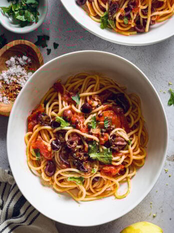 Plated spaghetti alla puttanesca topped with olives, capers, parsley, and lemon zest, with flake salt and chili flakes on the side.