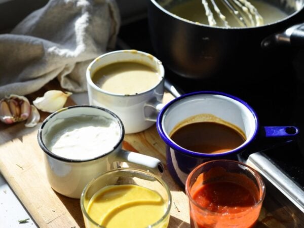 five mother sauces in mismatched cups on a wooden board by a sunlit kitchen window with a whisked pot and scattered garlic
