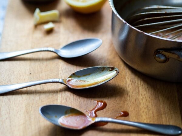 three tasting spoons with uneven sauce smears on a wooden board beside a lemon, salt, and a used saucepan in soft window light