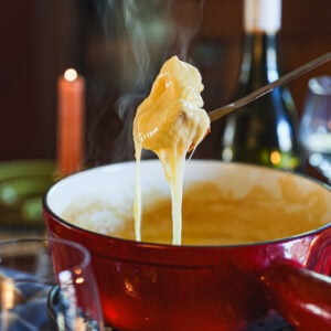 Melted cheese stretches from a fondue fork lifting bread over a steaming red pot, with wine glasses and a bottle in the background.