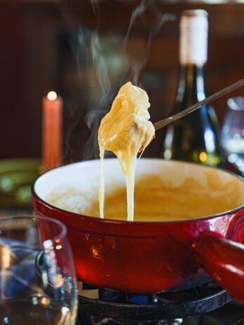 Melted cheese stretches from a fondue fork lifting bread over a steaming red pot, with wine glasses and a bottle in the background.
