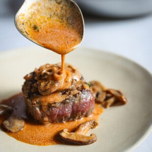 Spoon drizzling creamy mushroom Madeira sauce over a seared filet mignon plated with mushrooms, skillet of sauce in the background.